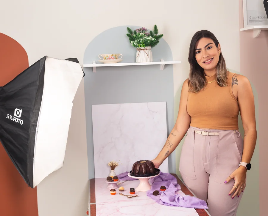 Lorena Moura ao lado do cenário montado para fotografar um bolo, com softbox de iluminação e prateleira decorativa